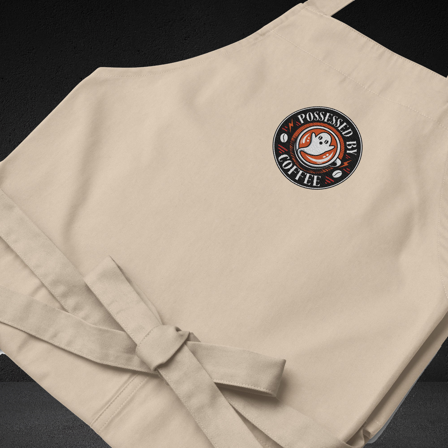 A beige apron with a circular logo on the front, featuring a ghost and the text "POSSESSED BY COFFEE".