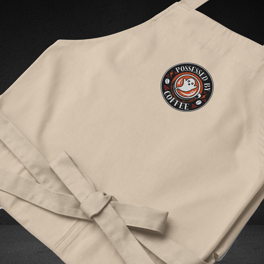 A beige apron with a circular logo on the front, featuring a ghost and the text "POSSESSED BY COFFEE".